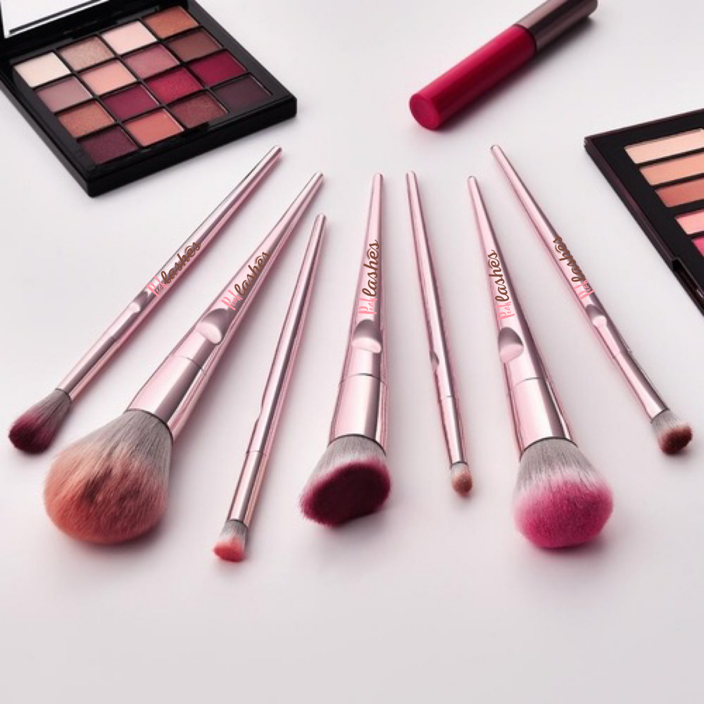 Set of makeup brushes with pink handles and varying bristle sizes, surrounded by makeup palettes and a lip gloss on a light background.